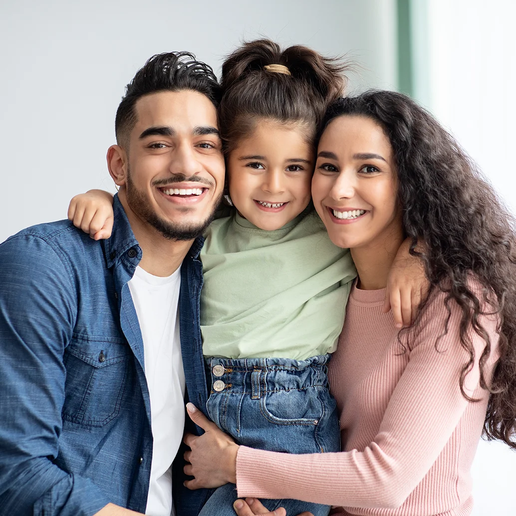 Familia de tres con hija, sonriendo abrazados frente a la cámara.