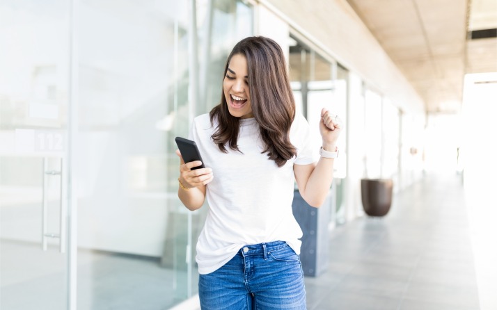 Mujer caminando sobre acera, sonriendo feliz mientras ve la pantalla de su celular.